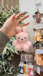 A hand holds a pink, plush Miffy bunny keychain with a gold keyring. Miffy merchandise and a price tag are visible in the background.