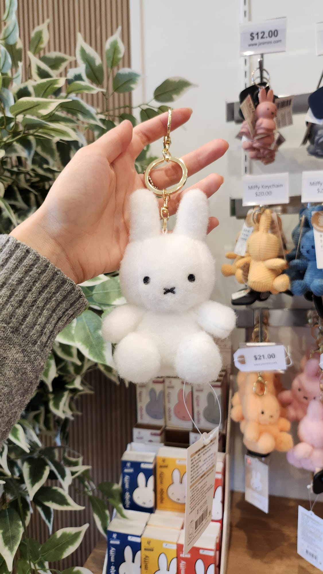 Close-up of a hand holding a fluffy white Miffy keychain with a gold ring. Various Miffy keychains and small Miffy boxes are visible in the background.