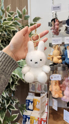 Close-up of a hand holding a fluffy white Miffy keychain with a gold ring. Various Miffy keychains and small Miffy boxes are visible in the background.