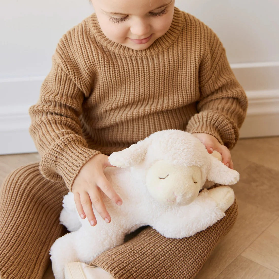 Child in a brown outfit holding a white Labmie plush toy on a light wooden floor.