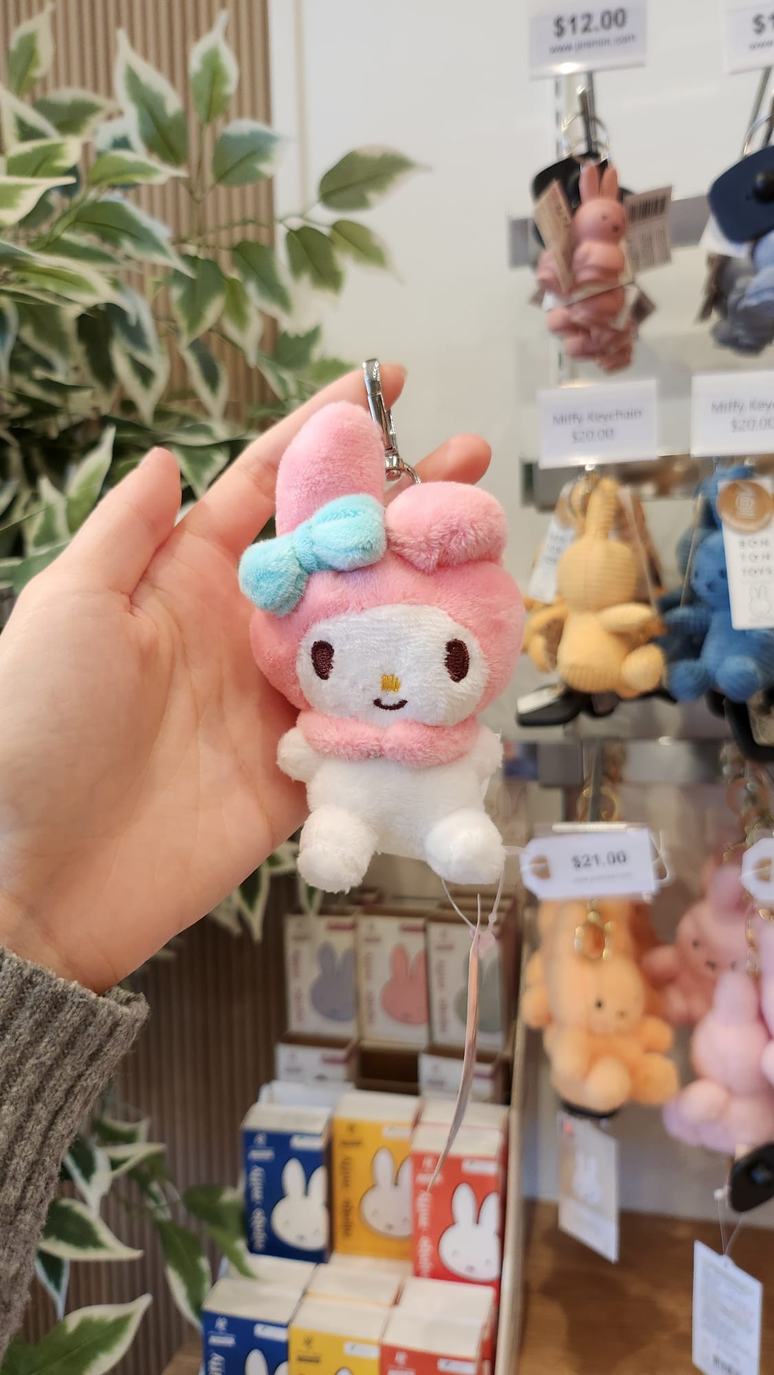 Close-up of a My Melody plush keychain held by a hand in a store setting, surrounded by other keychains and product packaging.