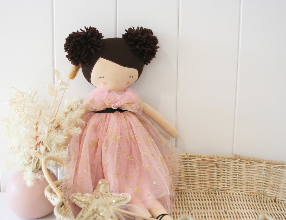 A doll with brown hair in buns wearing a pink dress with gold stars sitting next to a woven basket and a pink vase with dried flowers.