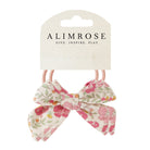 Two hair bows with pink elastic bands, displayed on a cardboard tag with the brand name "Alimrose." The bows are floral printed with pink and green on a cream background.