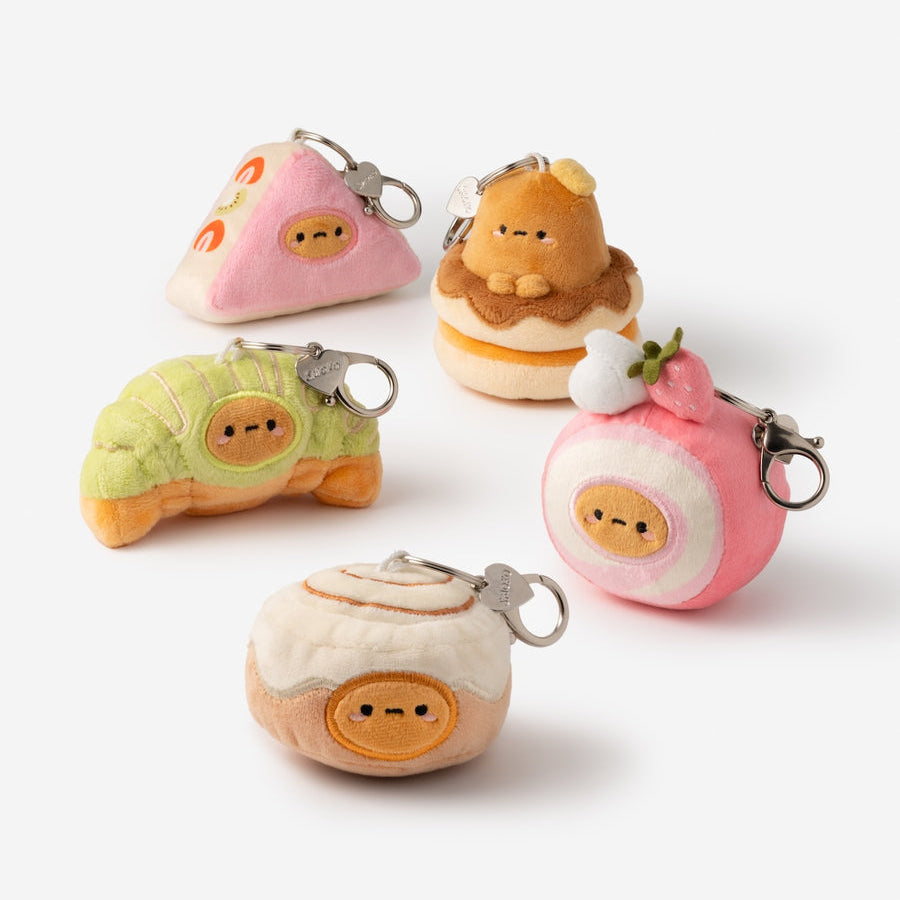 Five cute plush food-themed keychains arranged on a clean white background. The keychains include a pink slice of cake, a brown cookie on a cupcake, a green croissant, a pink strawberry pastry, and a cream-colored dumpling with orange facial features.