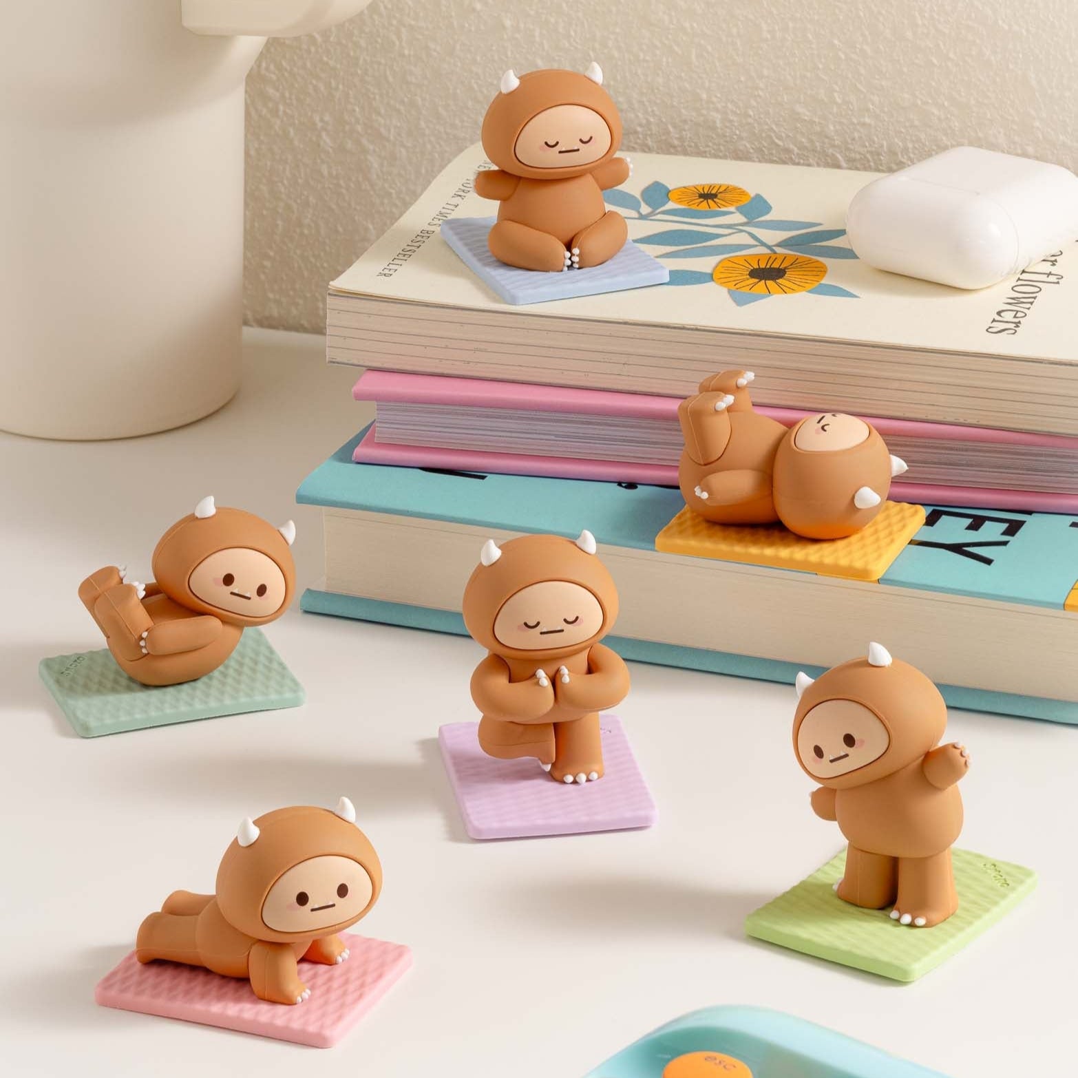 Collection of yoga-posing brown figurines on colorful mats, stacked books, and a white AirPod case.