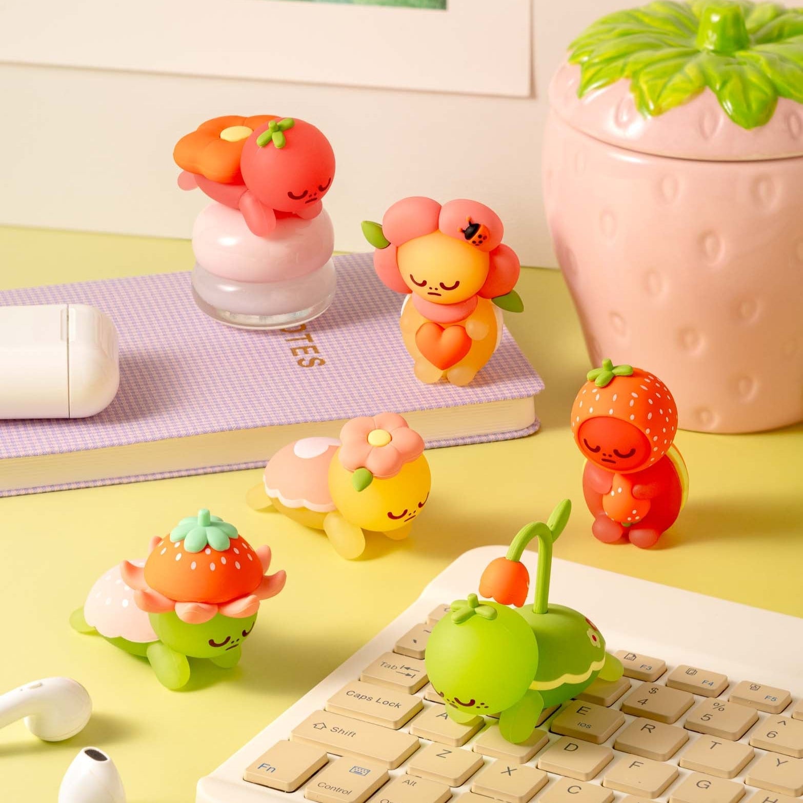 A yellow desk with a lavender notepad, white wireless earbuds and keyboard, and several small colorful fruit-themed figurines.