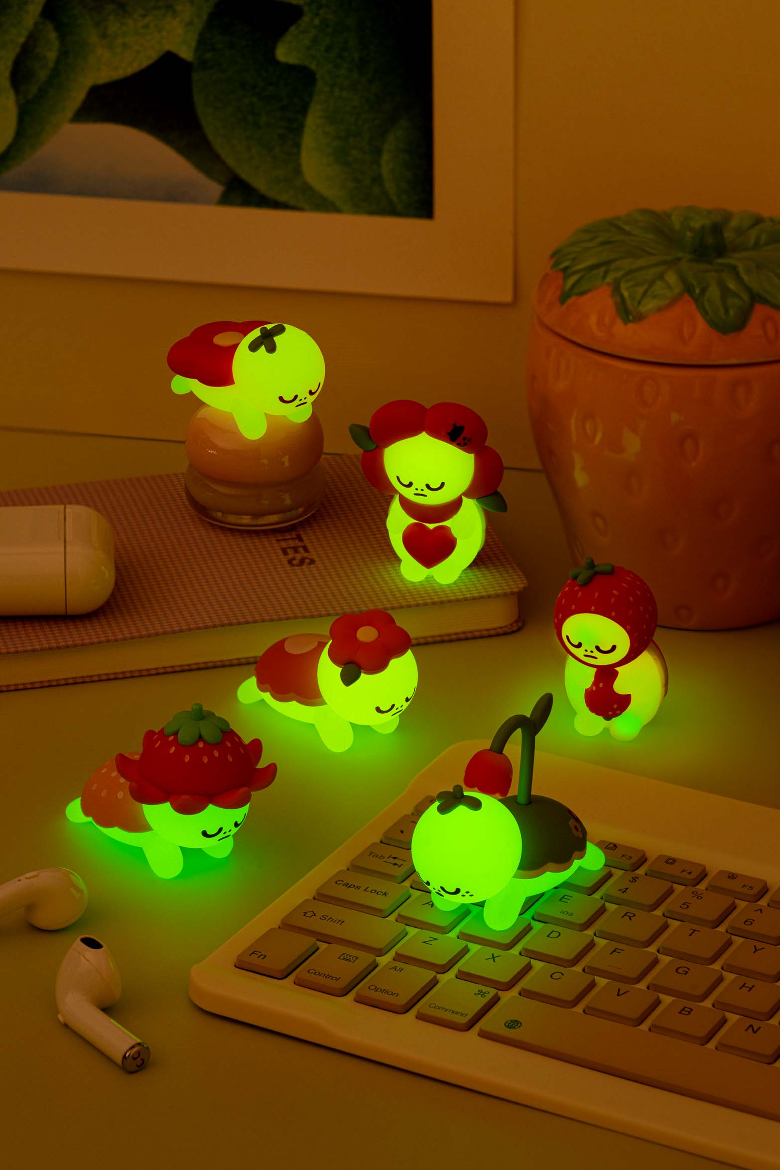 Six small, glowing, cartoon-like figurines in the shape of animals and fruits arranged on a desk with a yellow keyboard, a notepad, and wireless earphones.