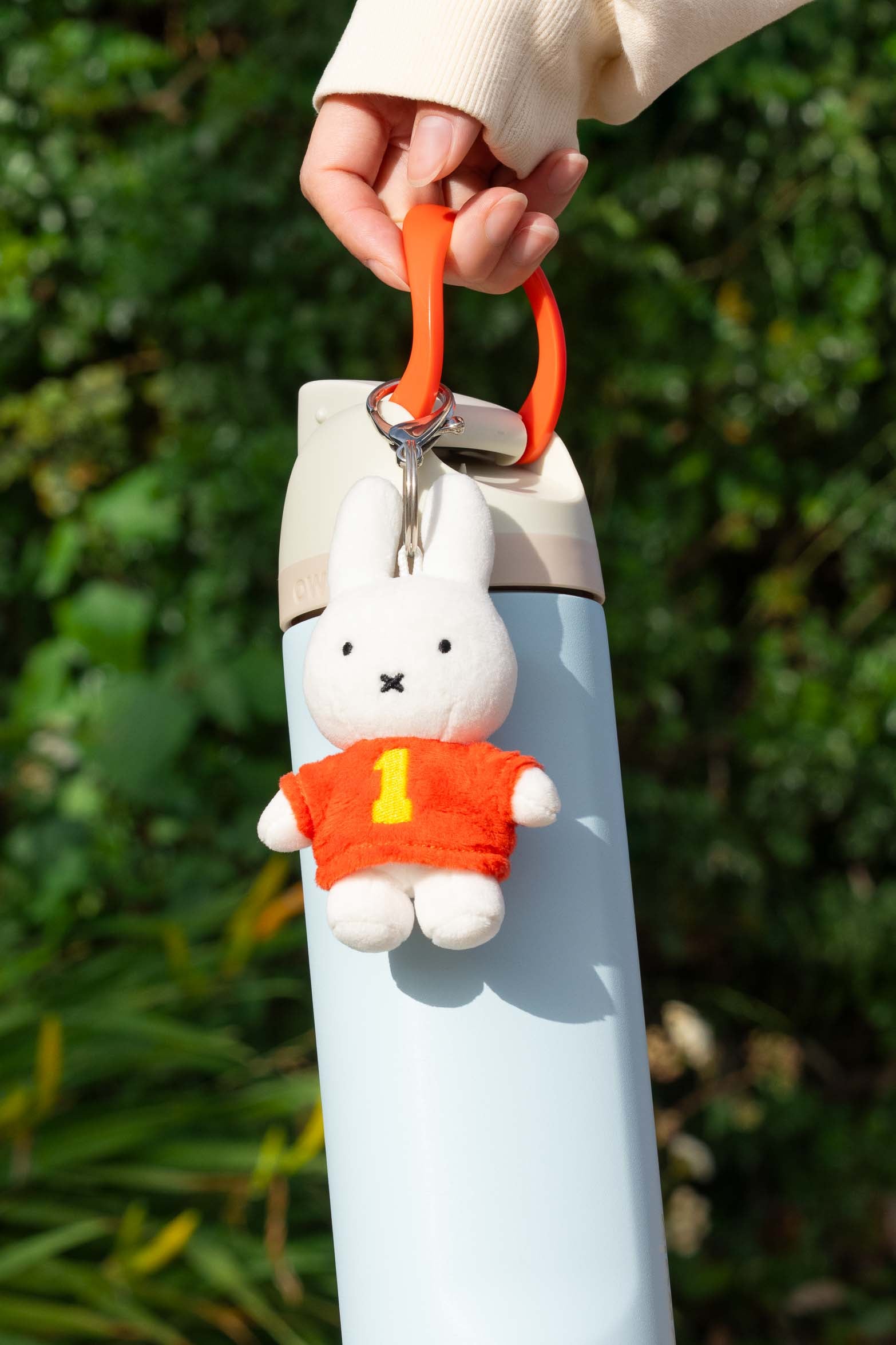 Person holding blue water bottle with Miffy plush keychain with orange shirt and the number one.