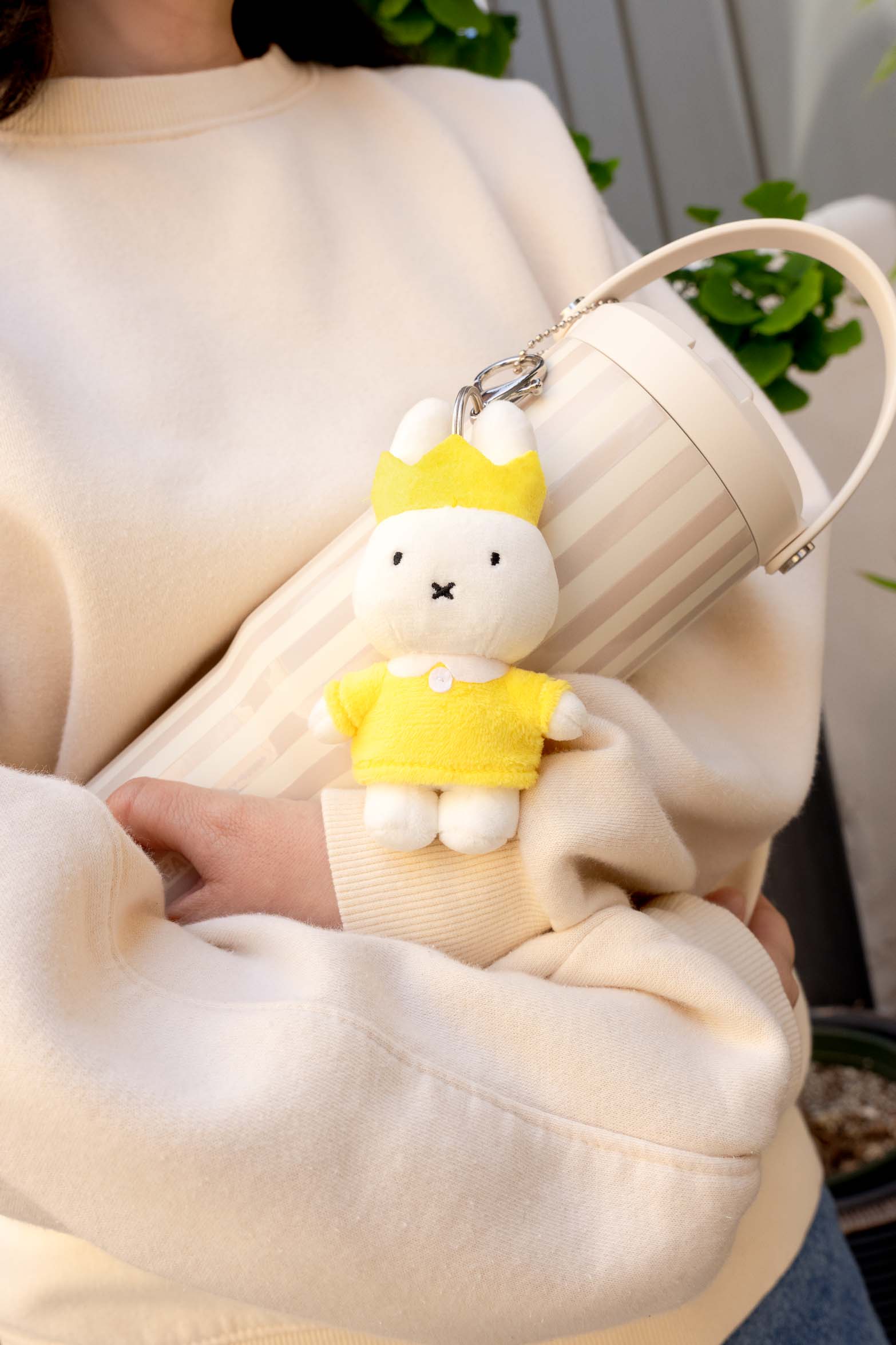 A woman wearing an off-white sweatshirt is holding a striped cylindrical container with a Miffy keychain.