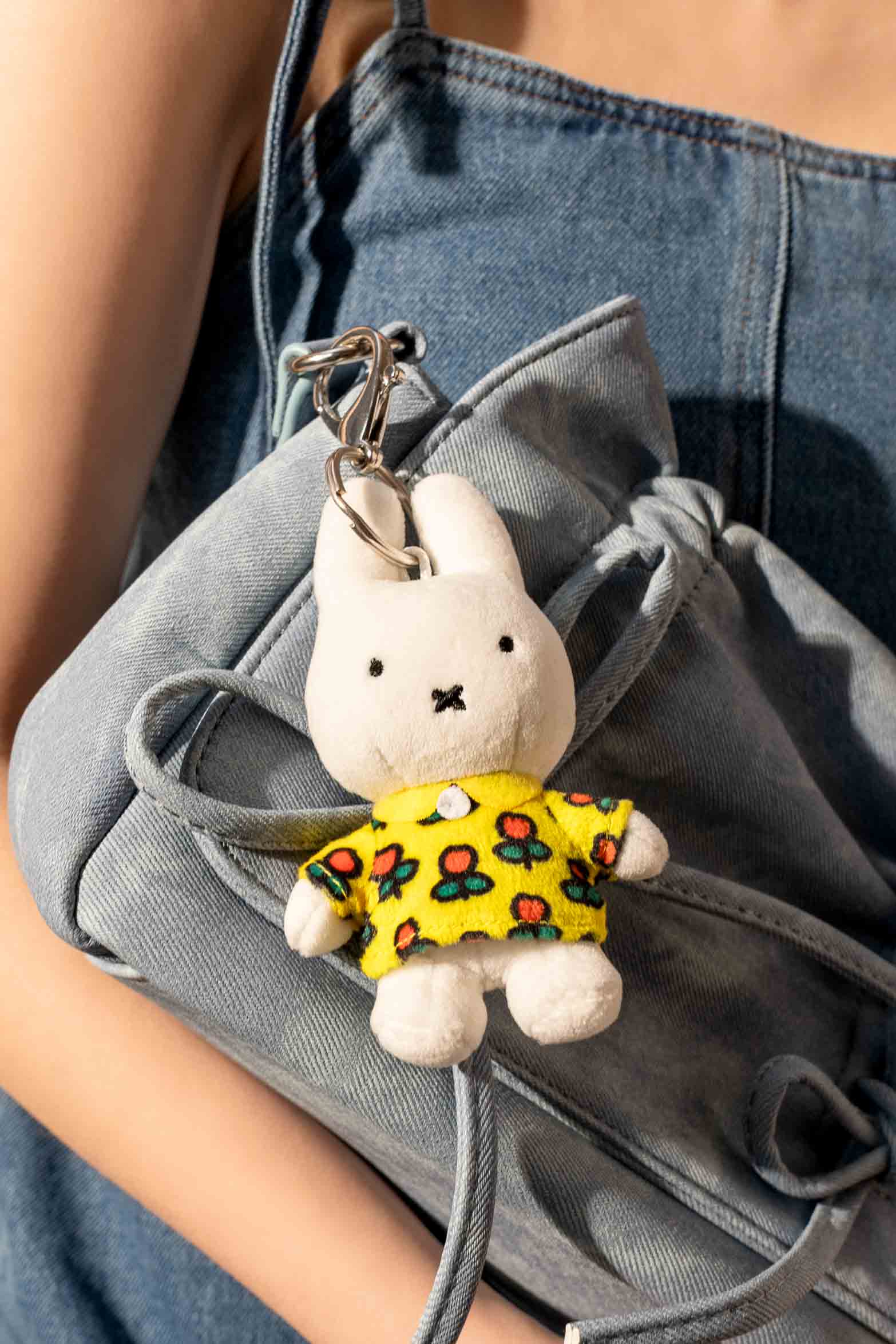 Miffy plush keychain with a yellow floral shirt, attached to a denim drawstring bag, held by a person in a denim outfit.