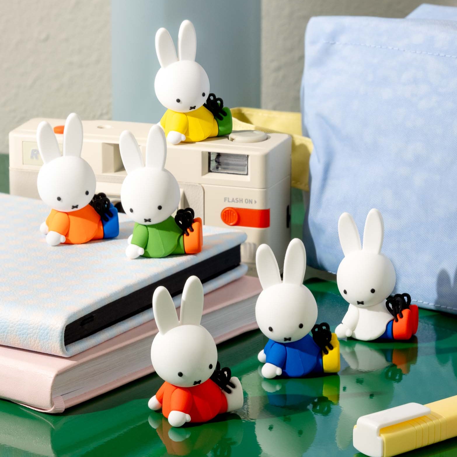 Six white Miffy bunny figurines in red, orange, green, yellow, and blue clothing, holding small black items, arranged on a green surface with notebooks, a vintage camera, a blue pillow, and a highlighter.