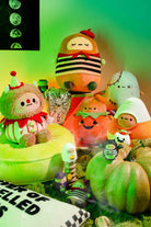 Assorted Halloween-themed plush dolls, including a clown, pumpkin, candy corn, and a zombie, arranged on a green cushion and among green pumpkins.
