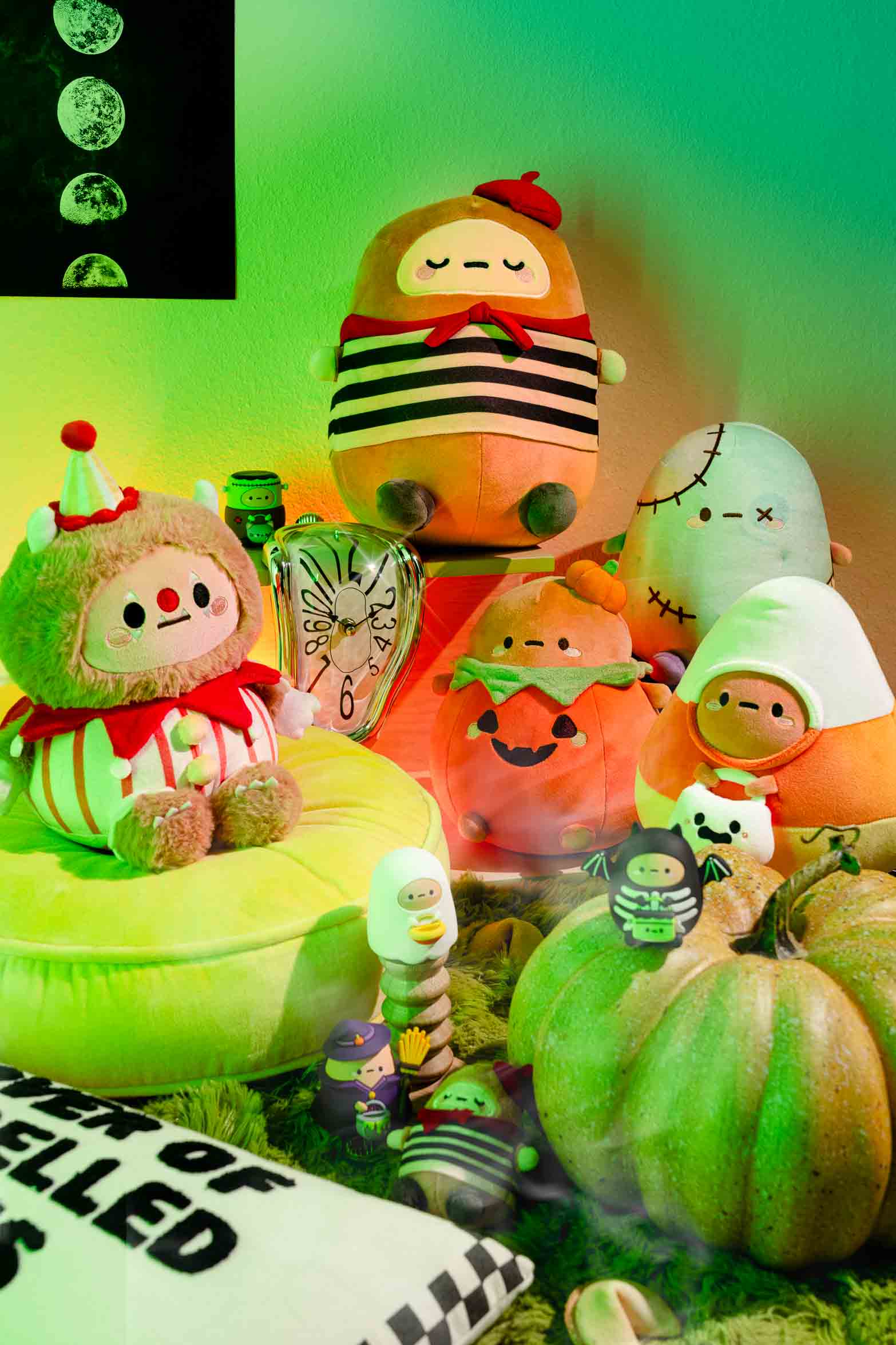 Assorted Halloween-themed plush dolls, including a clown, pumpkin, candy corn, and a zombie, arranged on a green cushion and among green pumpkins.