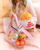 A person in a pink ribbed shirt and white skirt sitting on a peach-colored rug, holding a pink bag with novelty plush keychains and a plush doll wearing a shirt that says "But Daddy I Love Him."