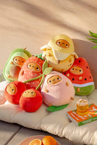 Group of fruit-shaped plush toys with faces on a white cushion, beside a glass of iced tea and a book.