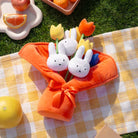 A bouquet of stuffed Miffy bunnies and tulips wrapped in orange fabric with a bow, resting on a yellow and white checkered blanket.