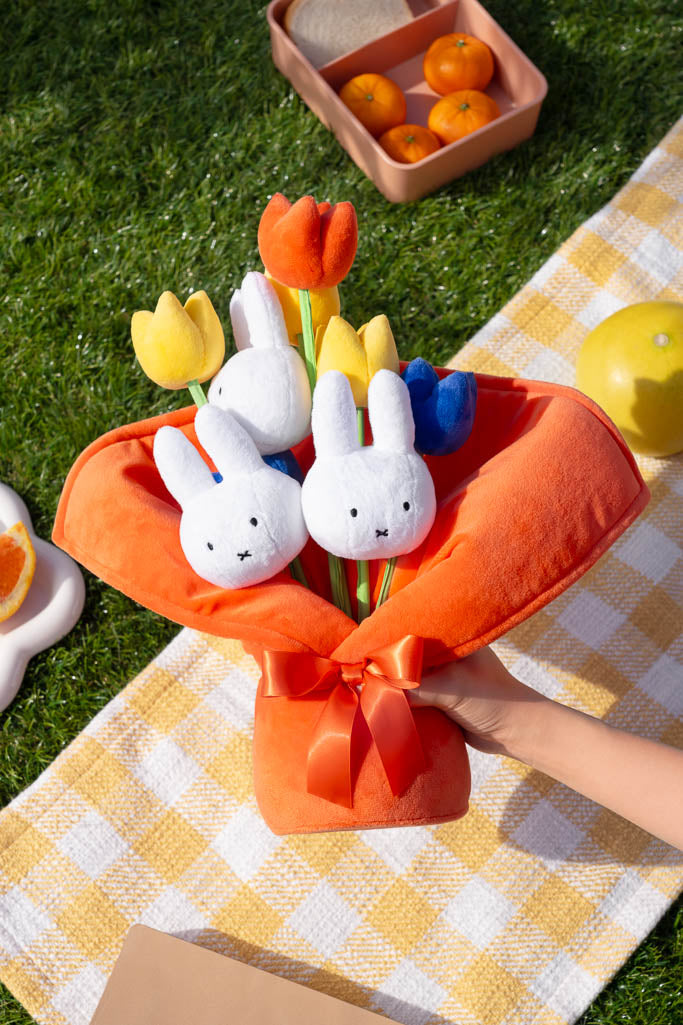A bouquet of plush bunny and tulip toys wrapped in orange fabric held over a yellow checkered blanket on grass.