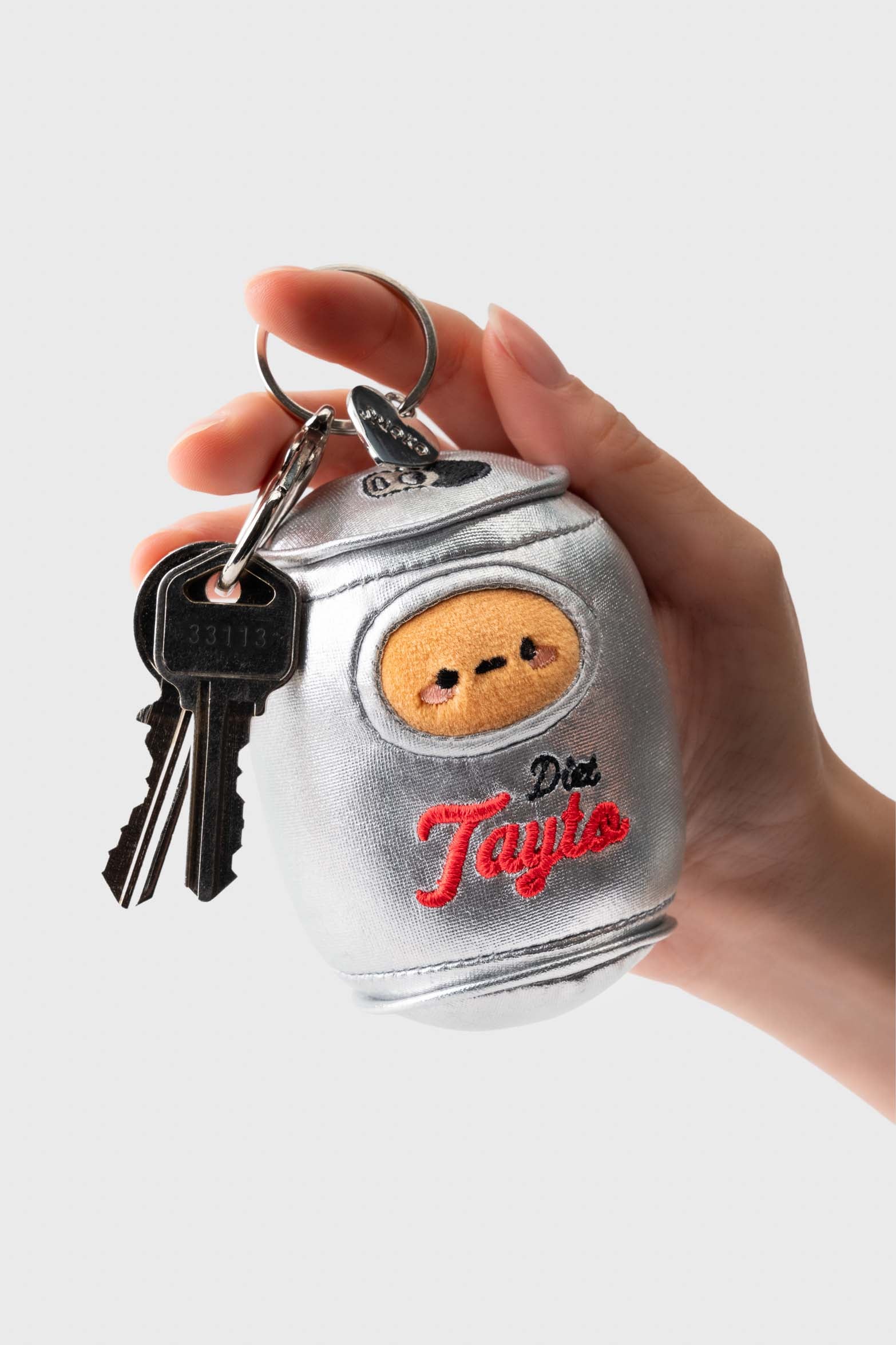 Hand holding a silver, plush keychain in the shape of a soda can, with keys attached. The can has a cartoon face and embroidered red script that reads "Diet Tayto."