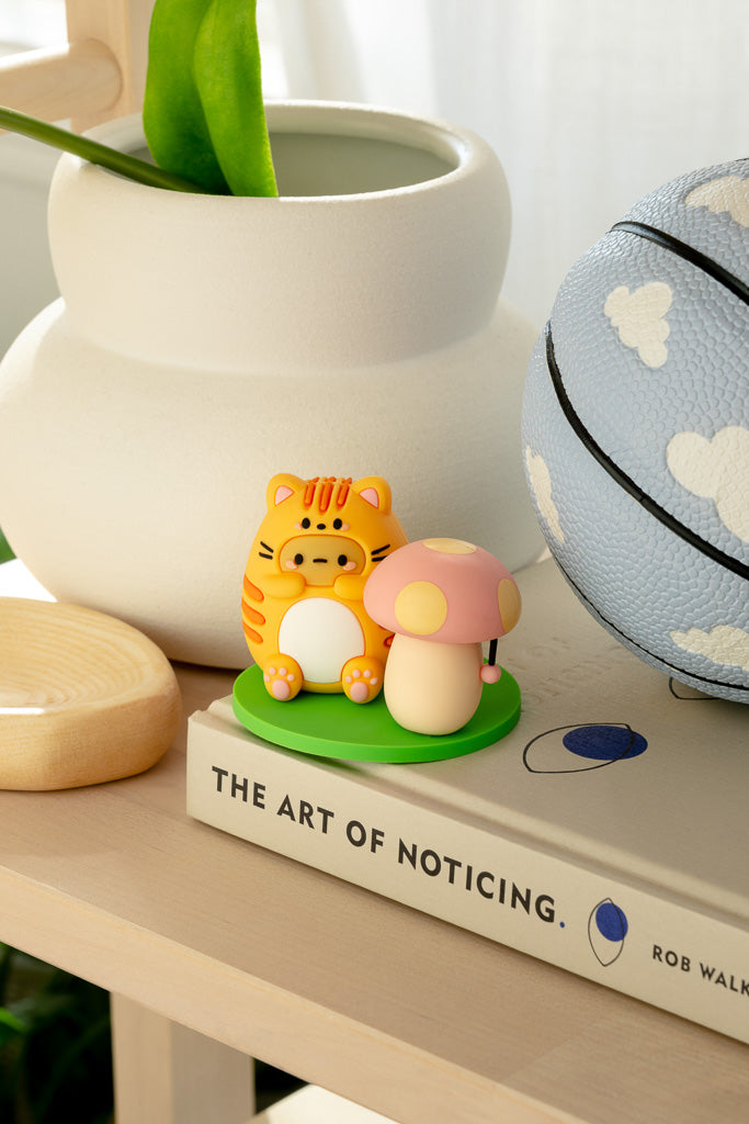 A still life featuring an orange tiger figurine, a pink mushroom figurine, a light blue basketball with white clouds, and a white vase, all on a book titled "The Art of Noticing."