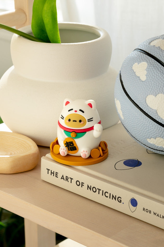 Close-up shot of a lucky cat figurine on a book titled "The Art of Noticing" with a white vase and blue basketball in the background.
