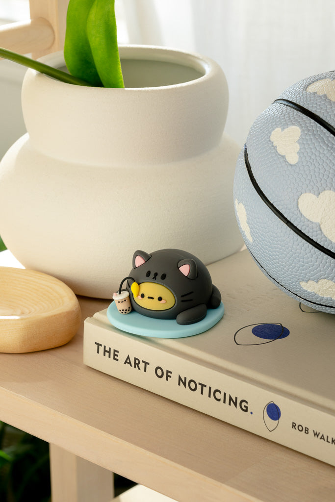 Cat figurine holding bubble tea on "The Art of Noticing" book, next to a blue basketball with white cloud design.