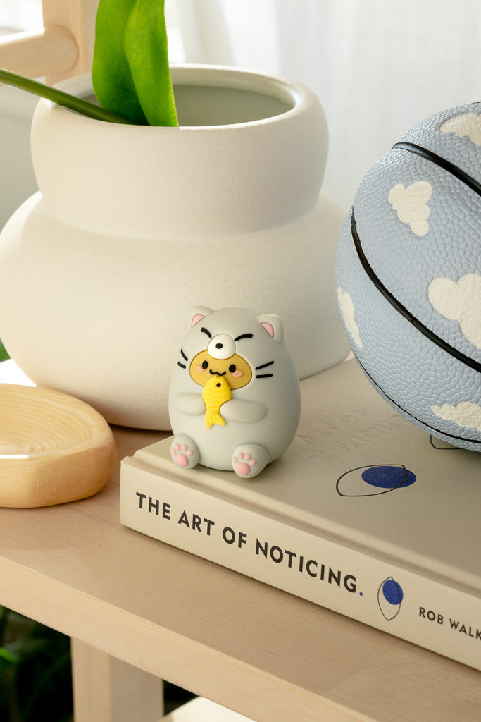A gray cat figurine holding a yellow fish sits atop a book next to a white vase and blue basketball.
