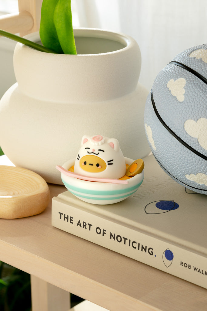 Close-up of a light tan shelf with a white vase, a wooden coaster, a figurine of a pig in a bowl of ramen on top of a book called "The Art of Noticing," and a light blue basketball with cloud designs.