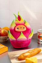 A plush, anthropomorphic dragon fruit sits on a kitchen counter surrounded by food items and cooking accessories.