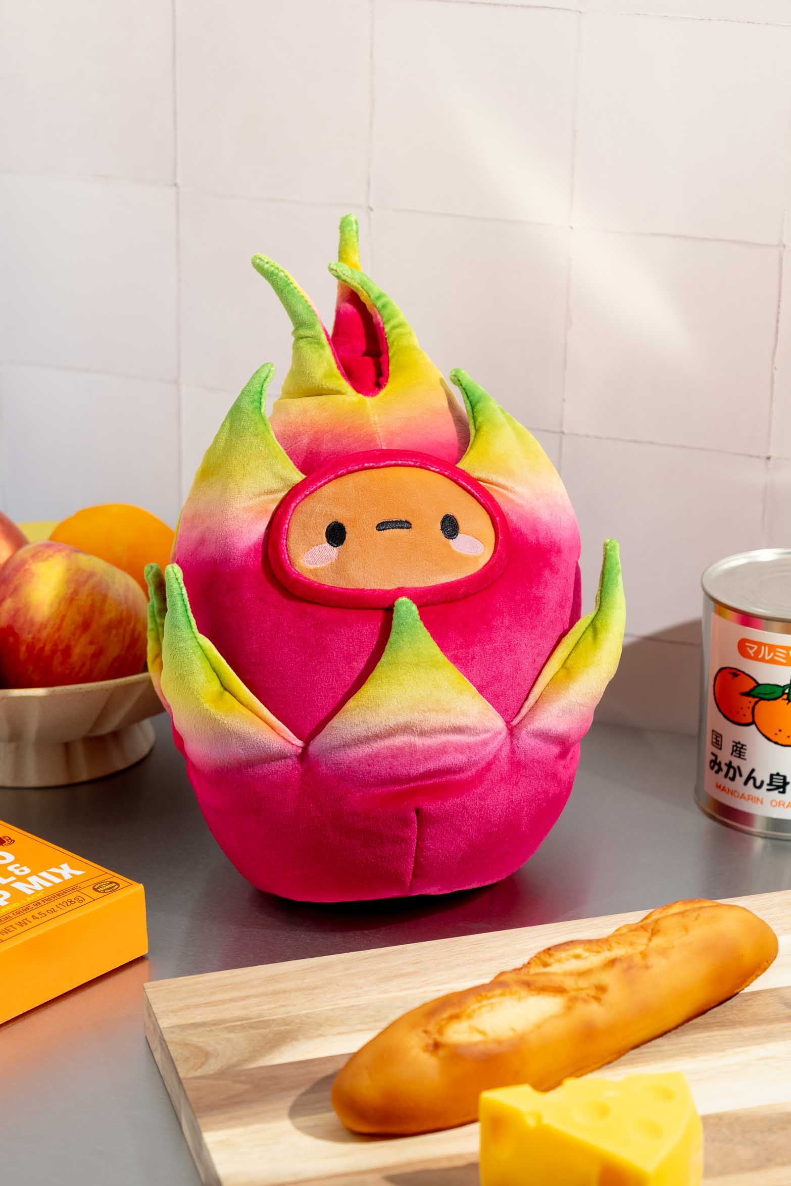 A plush, anthropomorphic dragon fruit sits on a kitchen counter surrounded by food items and cooking accessories.
