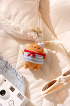 Still life with a plush sailor potato keychain, a white corduroy bag, a white point-and-shoot camera, light-wash denim, and cream sunglasses on a cream quilted blanket.