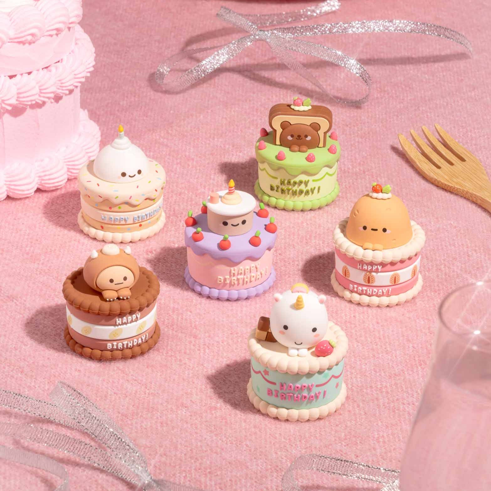 Six miniature cakes with cartoon characters on top, arranged on a pink textured surface with ribbons and a wooden fork.