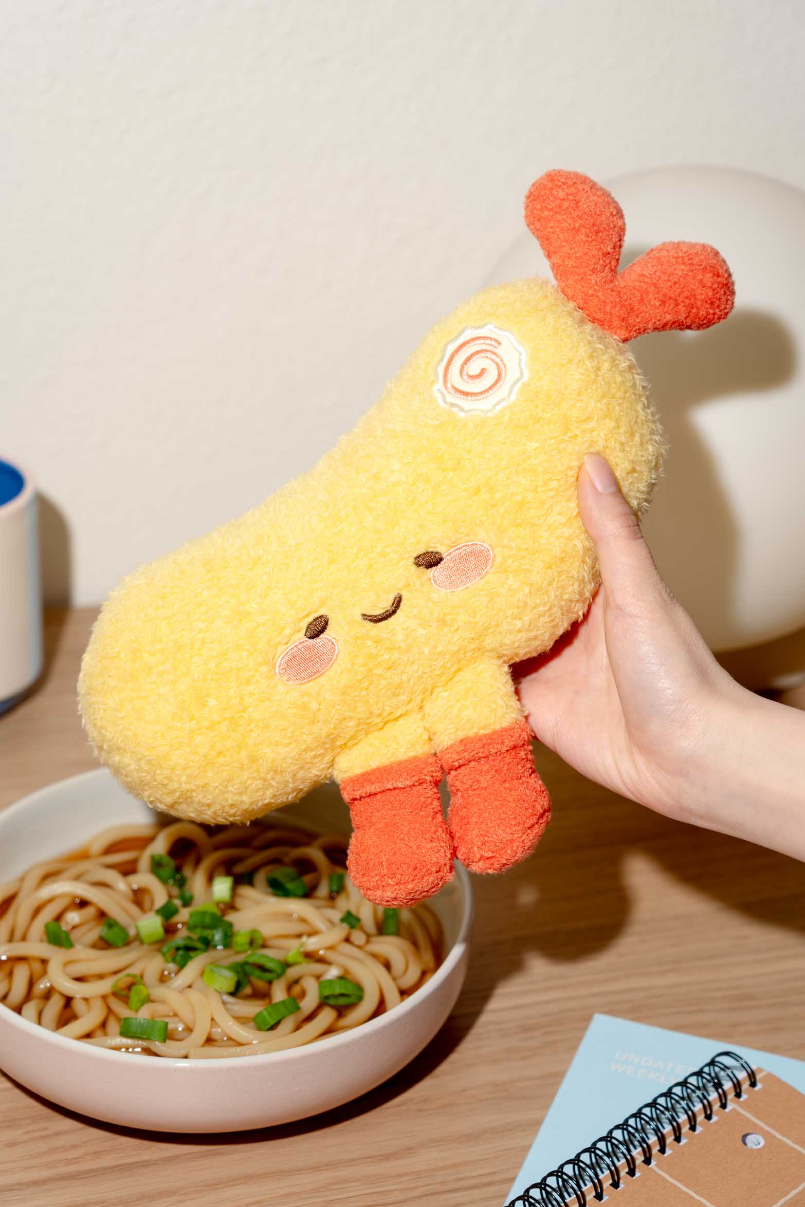 Close-up of a yellow tempura plush toy with orange accents, held in a hand above a bowl of noodles.