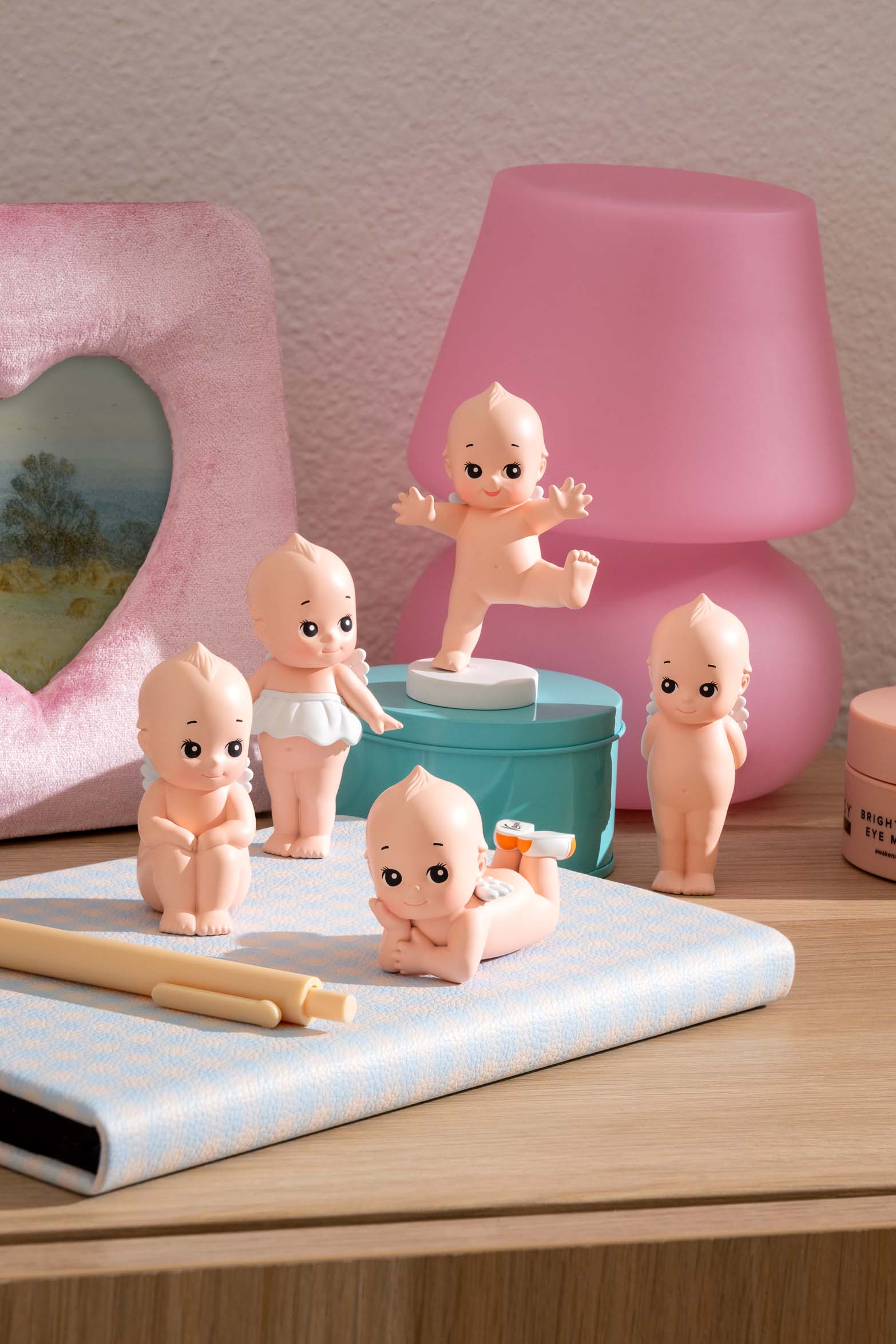 Five naked Kewpie dolls, turquoise storage box, pink heart frame, light blue notebook, and pink lamp on a wooden surface.