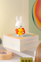 White rabbit figurine holding a yellow teddy bear on a colorful block with a rainbow wall in the background.