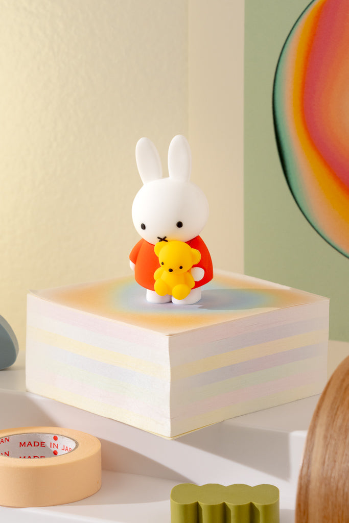 White rabbit figurine holding a yellow teddy bear on a colorful block with a rainbow wall in the background.