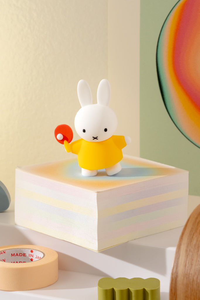 White rabbit figurine in a yellow outfit holding a red ball on a colorful block with a rainbow wall in the background.