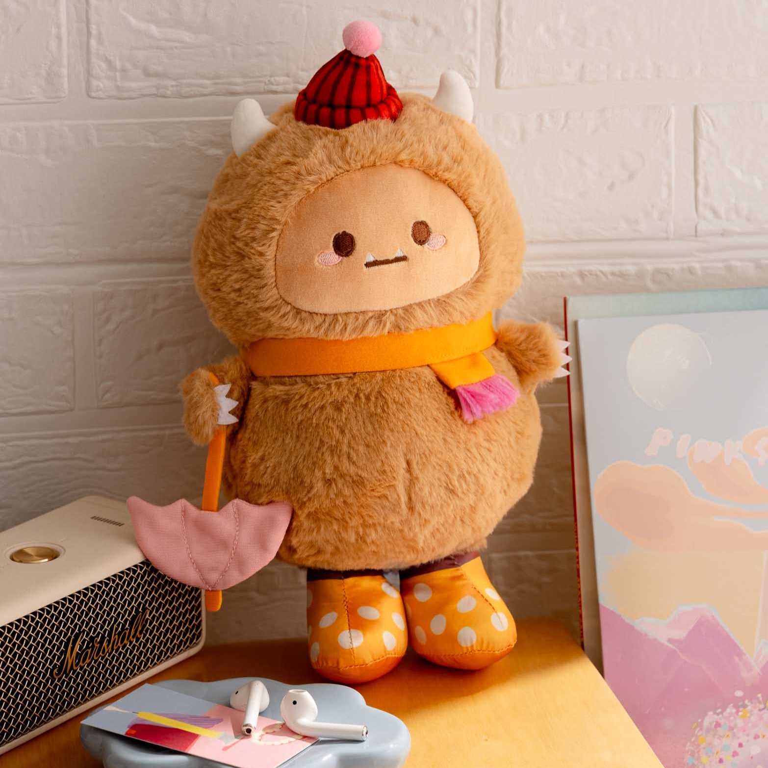Plush brown monster with a red hat, orange scarf, and yellow polka-dot boots holding a pink umbrella.