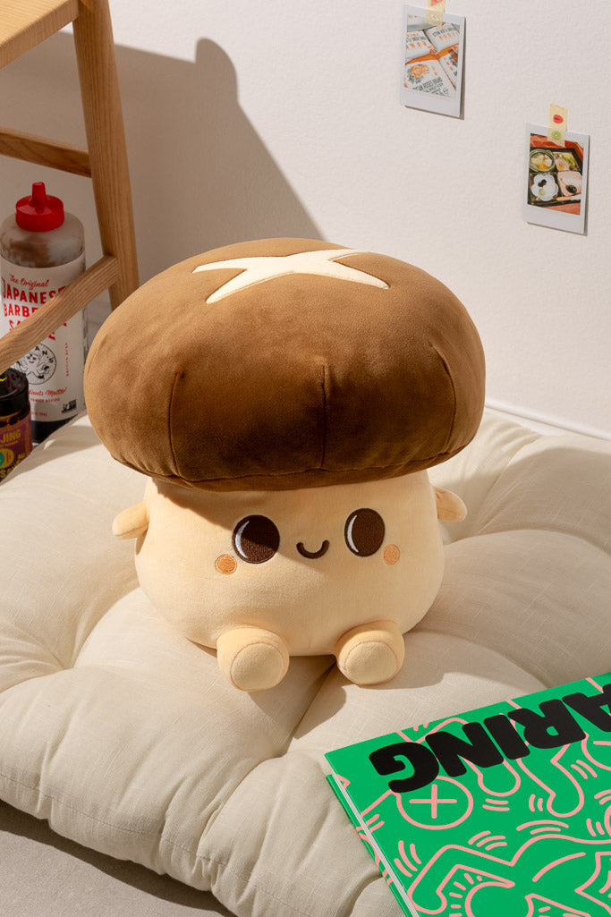 A mushroom-shaped plush toy with a brown cap and cream-colored body is resting on a beige cushion next to a green book.
