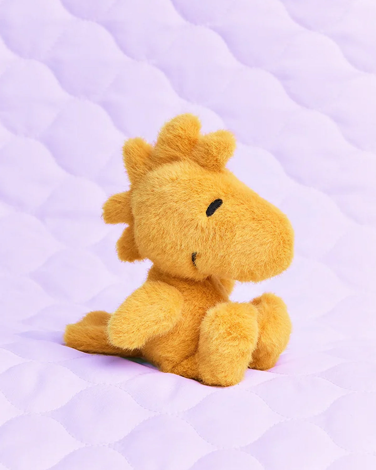 Orange, plush Woodstock toy sitting on a purple quilted surface.