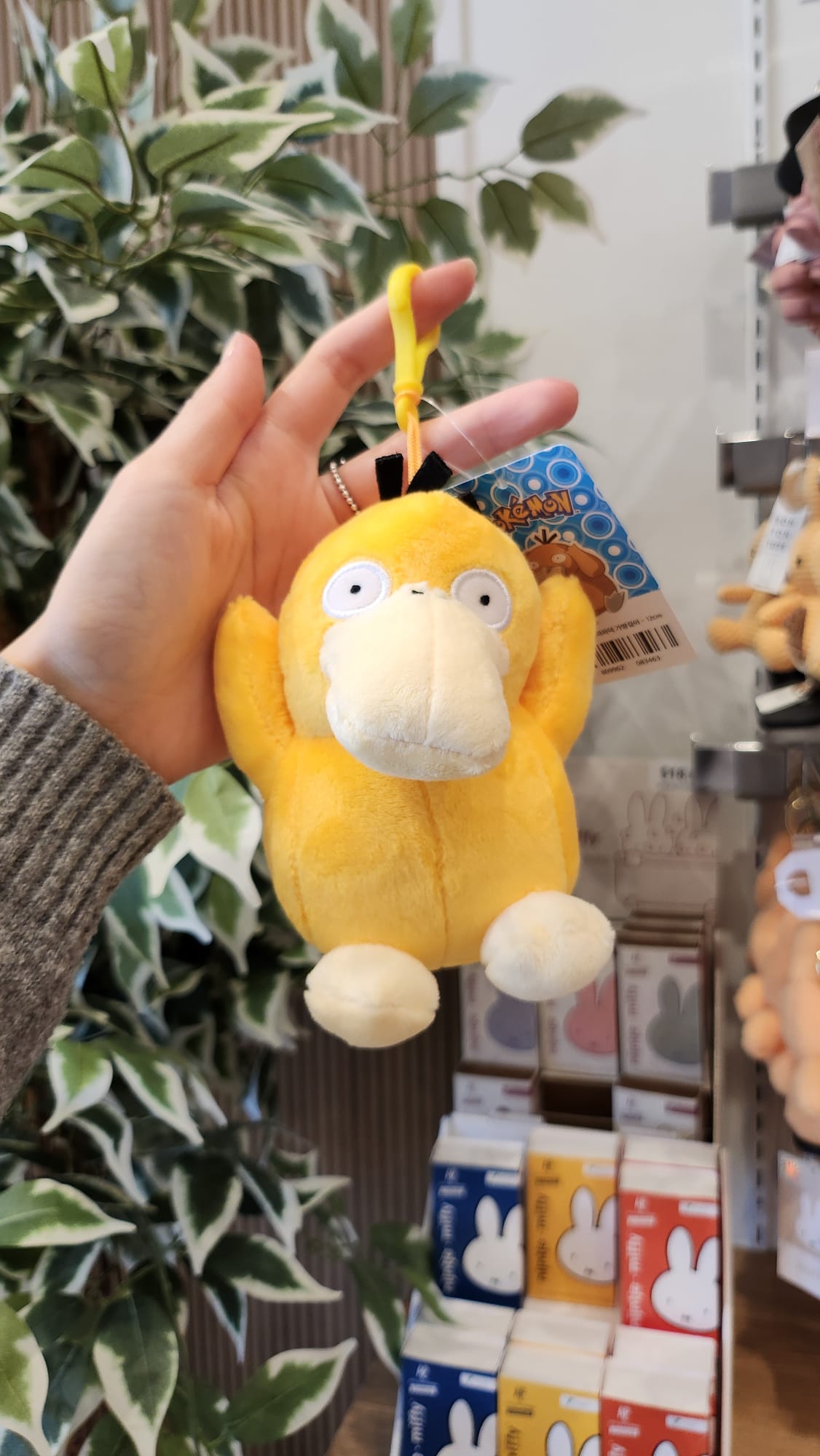 Close-up of a yellow Psyduck plush keychain held in a hand, with a colorful tag and retail displays in the background.