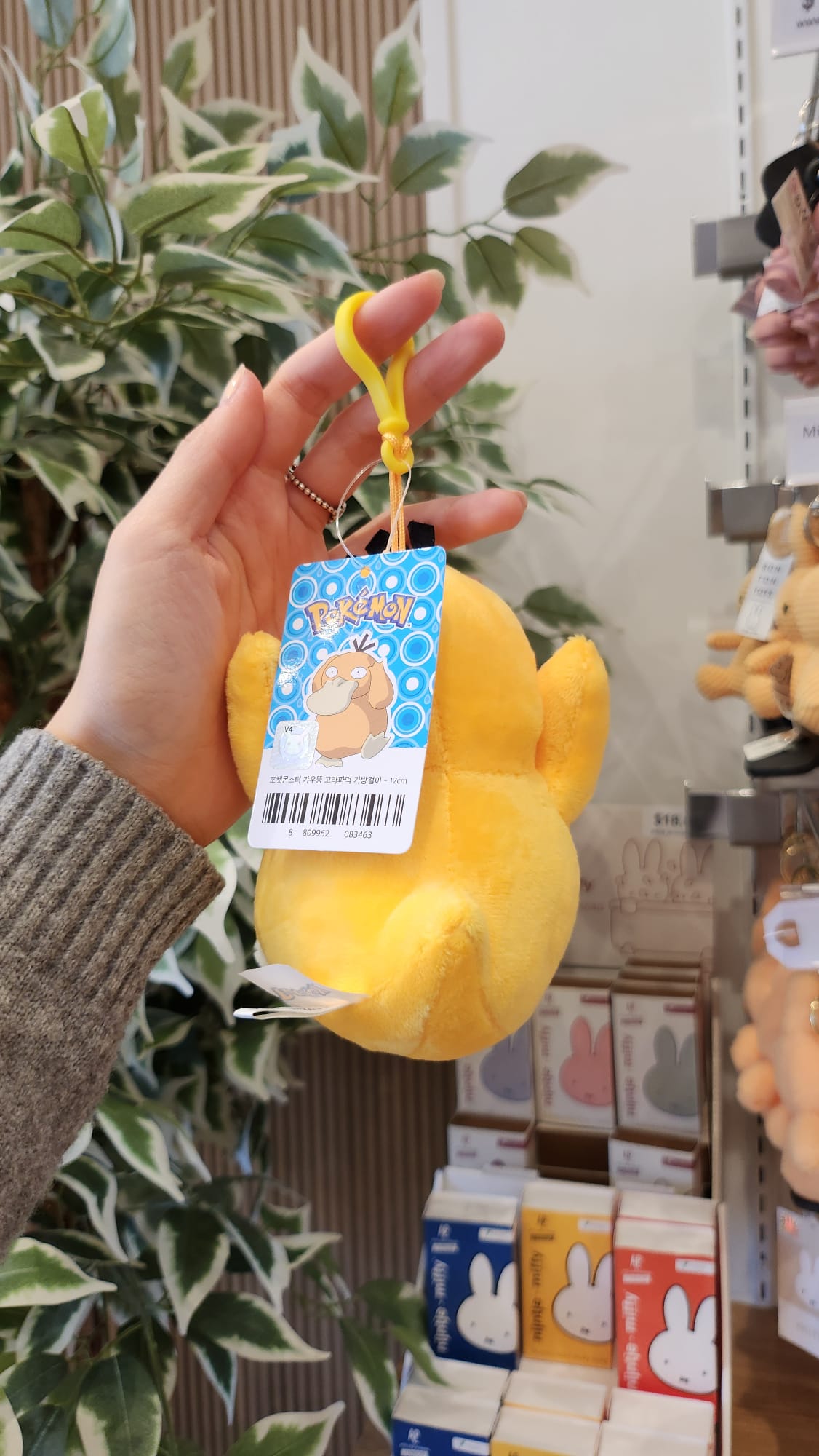Hand holding a yellow plush Psyduck keychain with a tag, near a plant and shelves of Miffy tissue boxes.