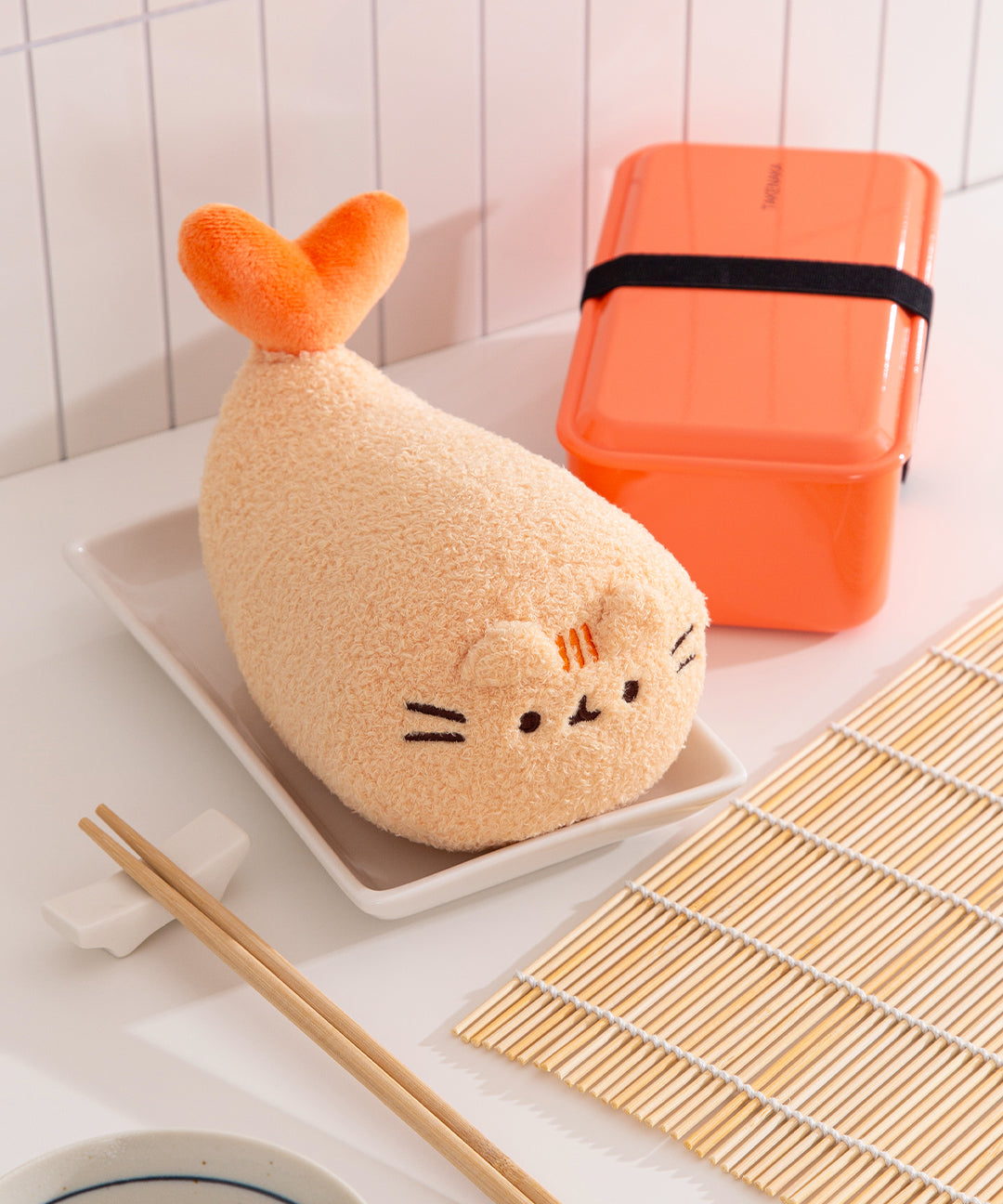 Plush toy shaped like a fish on a plate with an orange bento box and bamboo mat in the background.