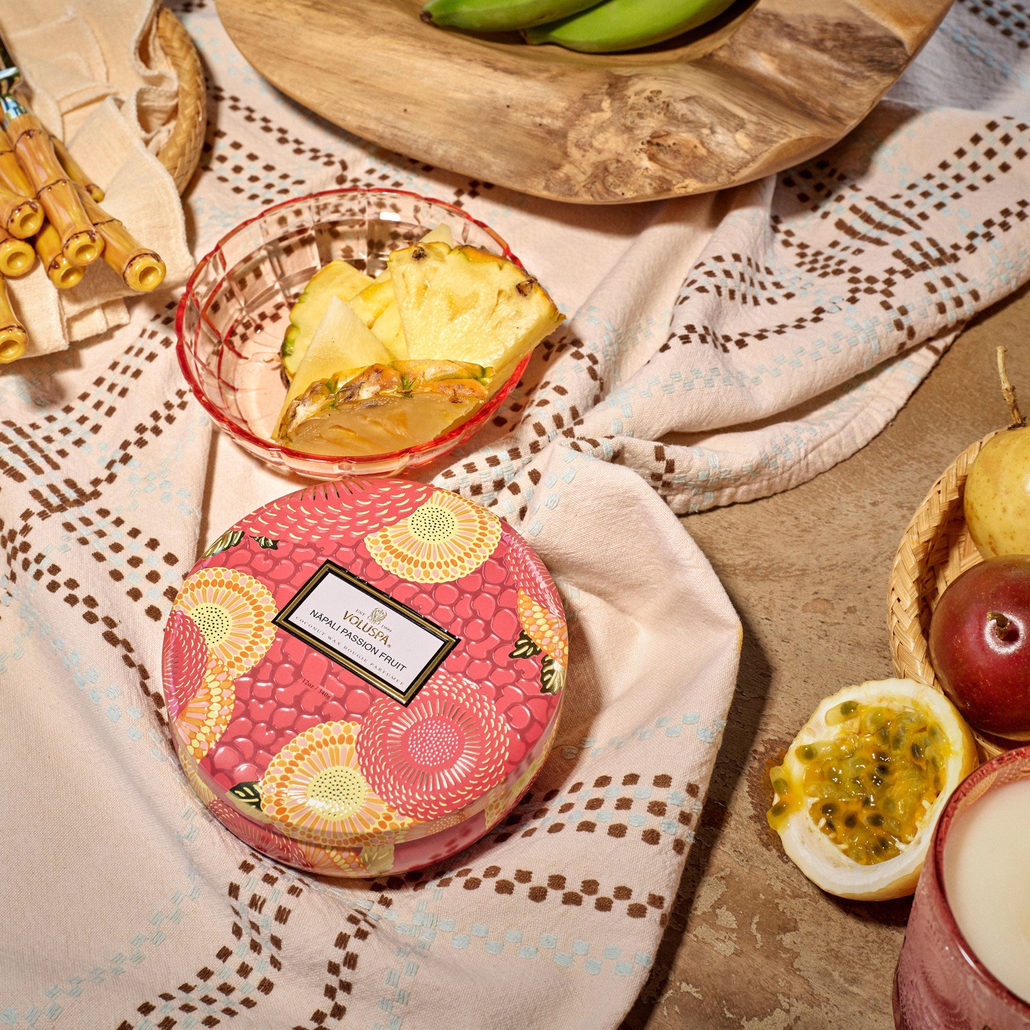 Decorative pink tin with fruit design on a patterned cloth with fruits and a bowl in the background.