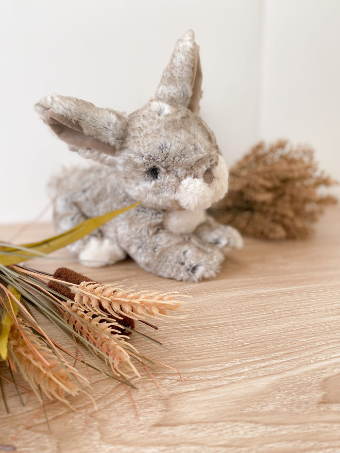 Stuffed gray rabbit with cattails and wheat on a light wooden surface.
