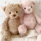 Two plush teddy bears, one brown and one pink, sitting on a white blanket.