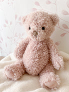 A pink, fuzzy teddy bear is sitting on a white blanket against a floral patterned wall.