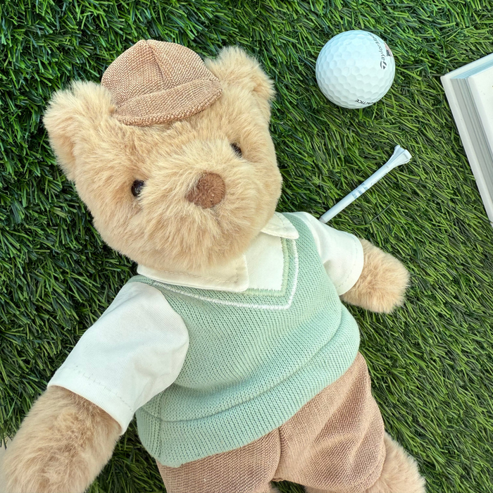 Teddy bear in a sweater vest and cap lying on grass with a golf ball and club nearby