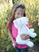Girl hugging white stuffed animal with pink ribbon in outdoor setting.