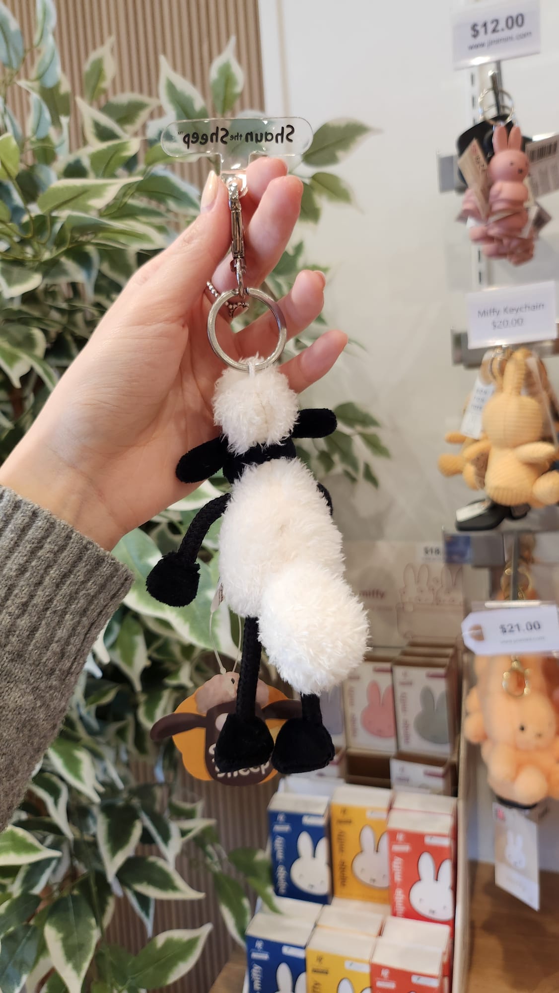 A hand holds a white and black sheep keychain in front of a shelf stocked with Miffy merchandise.
