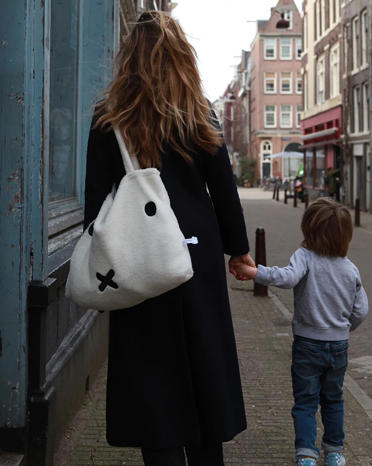 Woman in black coat with Miffy tote bag holding child's hand on a street.